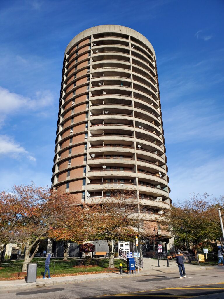 A round residential tower above a tree-lined sidewalk in Roxbury.